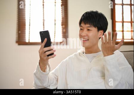 Happy young Asian man in casual clothes relaxes on a sofa at home, scrolling on his smartphone to read blogs or watch funny clips. A great lifestyle shot for living room, technology and online entertainment themes. Stock Photo