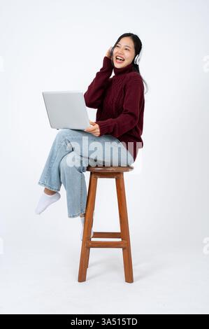 Cheerful young Asian woman in cosy clothes sits on a chair, wearing headphones and using a laptop. This relaxed indoor lifestyle shot captures a happy moment with music and tech, ideal for home office or casual leisure themes. Stock Photo