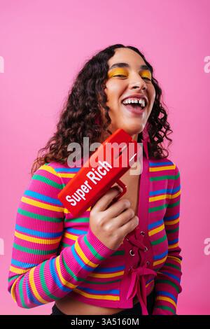Smiling Hispanic woman with a diastema holds a money gun in front of a pink studio backdrop. This playful, modern image suits entrepreneurship, influencer culture, and wealth or fashion campaigns. Stock Photo
