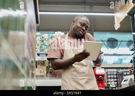 African American bartender using a tablet to place an online order while talking on the phone. The indoor cafe setting suggests hospitality tech and modern retail. Stock Photo