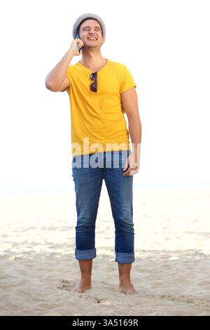 A cheerful Hispanic man wearing a hat stands barefoot on the beach, talking on his cellphone. The sunny seaside setting conveys a relaxed lifestyle and outdoor leisure. Ideal for travel, summer lifestyle, beach days, and mobile technology features. Stock Photo
