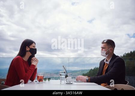 Group of casually dressed professionals outdoors at a restaurant, all wearing protective masks. They collaborate and brainstorm, illustrating teamwork in a modern business scene during the Covid-19 era. Stock Photo