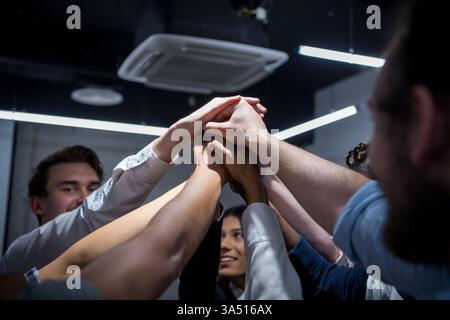 Diverse group of professionals in a modern office celebrate success with hands joined. They smile and stand together in a collaborative, corporate setting. Great for featuring teamwork, diversity and business achievement. Stock Photo