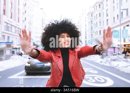 Black woman on a city street with an afro stands smiling as she makes a stop gesture. This expressive urban portrait communicates confidence and positive messaging for safety, awareness, or empowerment campaigns. Stock Photo