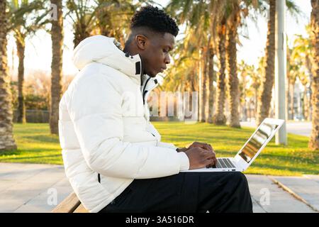 African American man sitting on a park bench working remotely on a laptop in sunny weather. This outdoor workspace evokes modern lifestyle and focus. Stock Photo