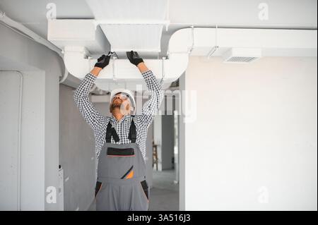 Indian male HVAC technician installing a ducted pipe system for ventilation in a residential building. The indoor workspace and professional equipment emphasize ductwork, heating, and air conditioning installation. Stock Photo