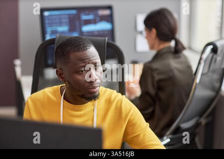 Portrait of a Black software developer at work in a modern office. The focused professional sits at a desk with a laptop, ready to code, with copy space to the side. A versatile image for tech industry, software development, and IT workplaces. Stock Photo