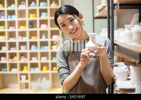 Chinese female potter showing pottery near shelves in a studio. The craftsperson appears proud of her work in a small business setting. Creativity, skill, and artistry are highlighted in this workshop scene. Stock Photo