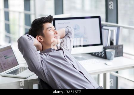 Smiling Chinese male office worker leans back with hands behind his head in a modern office. The relaxed pose signals confidence and job satisfaction in a professional setting. This casual moment reflects a positive workplace mood and focus on success. Stock Photo