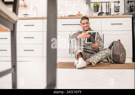 Smiling Arab military man in camouflage sits on the kitchen floor near a backpack, looking at a picture frame. This interior shot captures patriotism, family moments, and home life. Stock Photo