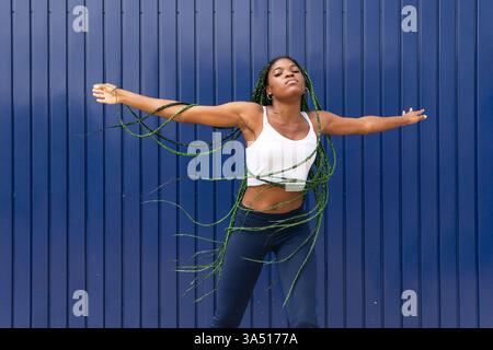 Cool African American woman dancing freestyle on a pavement, denim and white top with long braids against a blue metal wall. A dynamic urban lifestyle image capturing energy, movement, and street style for dance or fashion campaigns. Stock Photo