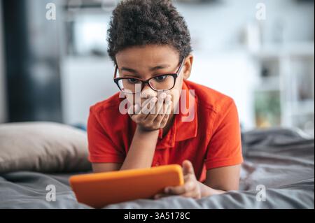 Curly-haired boy in an orange T-shirt engaging with content online on a digital device. A typical youth tech moment suitable for online learning, digital lifestyle, and connectivity topics. Stock Photo