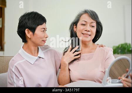 Smiling senior Asian woman sits on a sofa, admiring her makeup in a mirror after a beauty session with a middle-aged Asian friend. The moment highlights friendship, self-care, and cosmetic care at home in a cozy living room. Suitable for beauty, makeup, and aging gracefully themes in indoor settings. Stock Photo
