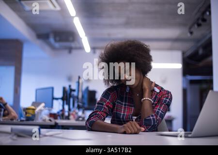 Smiling Black business woman with afro touching her neck getting tired from work while sitting at table with closed eyes in the office Stock Photo