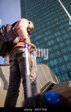 Portrait from behind of a young Black man ascending an airport escalator with a suitcase and smartphone in hand. The urban travel scene conveys movement and modernity, highlighting luggage and mobility. A versatile image for travel, commuting, and airport lifestyle campaigns. Stock Photo