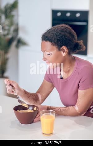 Mixed race woman with vitiligo enjoys a bowl of cereals at a kitchen table, highlighting breakfast time at home. This authentic lifestyle image focuses on wellbeing, skin positivity, and everyday nourishment. Stock Photo