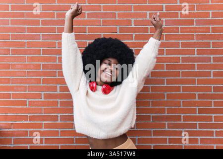 Vibrant lifestyle image of a cheerful African American woman dancing against a brick wall. She wears red headphones around her neck and sports an Afro hairstyle with a bright, white smile. Perfect for music, dance and lifestyle campaigns. Stock Photo
