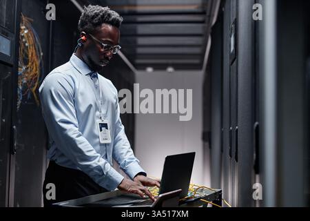 Backlit side portrait of a data engineer at work in a server room, using a laptop. Great for IT infrastructure, data centers, network engineering, and cloud computing topics. A professional, focused mood with a modern technology setting. Stock Photo