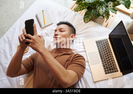 Young man lies on a bed at home, texting on his smartphone instead of coding on a laptop. The moment captures casual downtime, online connection, and the blend of work and leisure. Suitable for topics about social media use, remote work, and home office life. Stock Photo