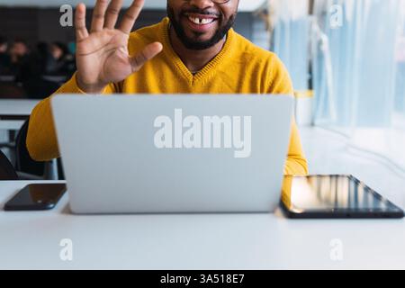 Anonymous happy Black man, a freelancer or student, waves to greet colleagues during a video conference on a laptop. This image suits remote work, online collaboration, and technology-driven communications. The upbeat mood and contemporary setup support content about virtual meetings and digital workplaces. Stock Photo