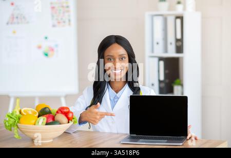 Positive female nutritionist pointing at a laptop with a blank screen in a medical office. The clean mockup style image includes copy space for website or marketing content. Ideal for nutrition, dietitian branding, and online health promotions. Stock Photo