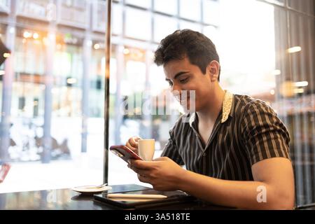 Smiling Hispanic man seated at a cafe table near a glass window. He holds a coffee cup and uses a smartphone, capturing a casual mobile moment. Great for lifestyle, cafe culture, and mobile technology topics. Stock Photo