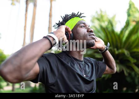 African American male athlete stands in a green tropical park with headphones, looking away. This fitness image suits sports, motivation, wellness and outdoor activity contexts. Stock Photo