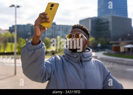 A Black man in a blue jacket taking a selfie in the city. Casual urban lifestyle image suitable for social media, technology and personal branding content. Stock Photo