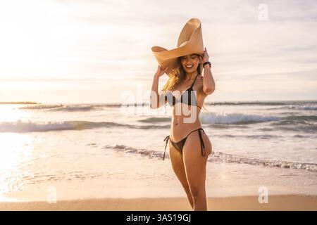 Cheerful Hispanic woman wearing black bikini looking down while standing with hand touching wide brim straw hat on beach on sunny day Stock Photo