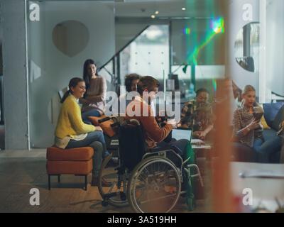 Inclusive business scene featuring a disabled man in a wheelchair participating in a team meeting in a modern open-plan coworking office. He wears virtual reality goggles while colleagues collaborate, with a drone providing assistance in the background. A forward-thinking image for accessibility, technology, and innovative work environments. Stock Photo