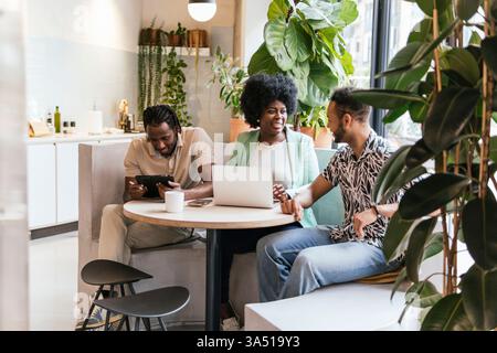 Three professionals from diverse backgrounds collaborate in a modern coworking space, with a laptop and tablet on the table. The group communicates, brainstorms, and shares ideas in a contemporary work environment. Perfect for business, team, and coworking branding shots. Stock Photo