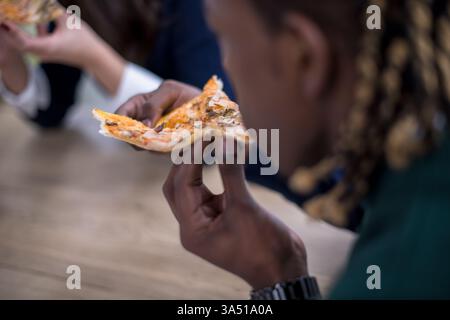 Black businessman eating pizza during a lunch break with colleagues in the office. A casual, diverse workplace moment ideal for corporate culture and team lunch themes. Stock Photo