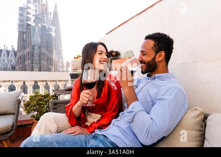 Couple on a rooftop balcony in Barcelona sharing an aperitif with city views near Sagrada Familia. A multiethnic, romantic scene ideal for travel, dating, and urban lifestyle campaigns. Stock Photo