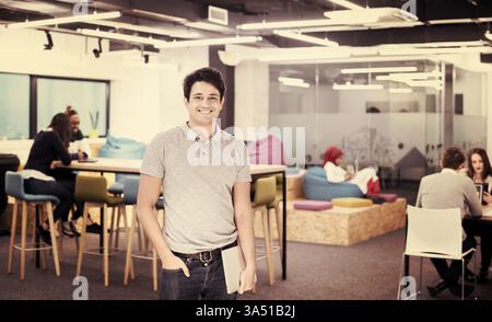 Smiling mixed race businessman holding a laptop, standing with his hand in his pocket beside colleagues in a busy office. This corporate portrait conveys leadership, teamwork and a modern workplace vibe. Stock Photo
