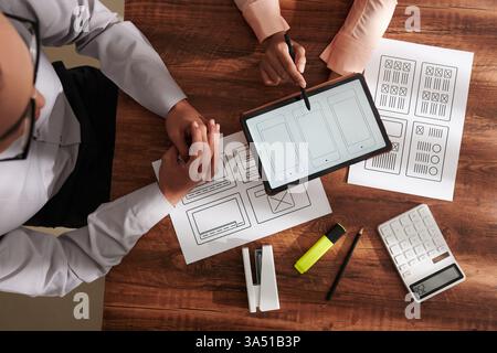 Top-down view of UI and UX designers collaborating on a mobile app interface during a meeting. The desk scene shows wireframes, tablets and documents as professionals brainstorm ideas. Great for technology, design, software development and teamwork contexts. Stock Photo