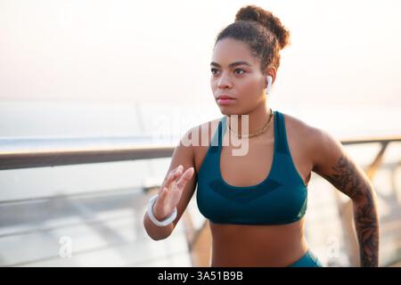 African American woman running outdoors in bright summer weather, listening to music with earphones. The image captures a healthy lifestyle moment during a solo jog. Perfect for fitness, training, and motivation content. Stock Photo