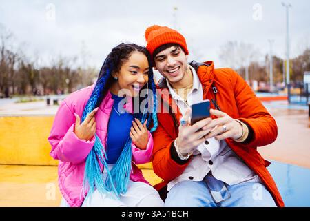 Multiracial young couple of lovers dating outdoors in winter, using a smartphone while wearing jackets. The scene conveys connection, social media, and seasonal style. Great for diversity, relationships, and mobile technology visuals. Stock Photo