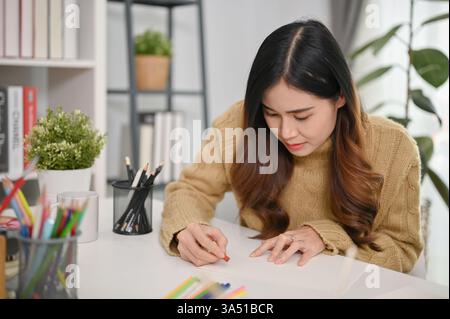 Focused Asian woman sitting at table painting on paper with oil pastel color at home Stock Photo