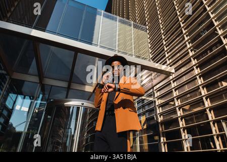 A Hispanic businessman wearing sunglasses and a fedora stands outside an office building, checking his watch while on a phone call. The urban setting and professional look suit business, corporate, and entrepreneur themes. Great for modern workplace campaigns. Stock Photo
