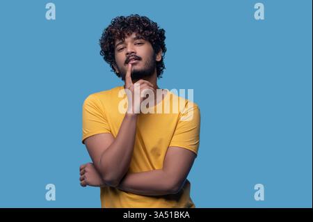 Portrait of a young Indian man with curly hair, standing with arms crossed and touching his lips with his index finger against a blue background. The pose communicates contemplation and confidence, suitable for business, tech, or lifestyle contexts. Ideal for corporate communications, marketing, or personal branding in studio settings. Stock Photo