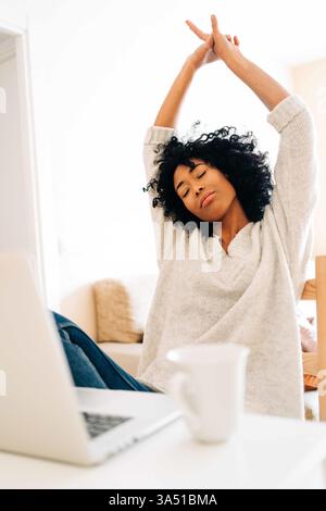 Black woman stretching arms while sitting at a table with a laptop at home. A relaxed remote-work moment that fits freelance, home office, and wellbeing themes. Stock Photo
