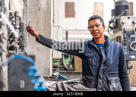 Smiling mixed race worker wearing safety glasses in a metal industrial factory standing among tools. He looks ready to work in a manufacturing environment, conveying professionalism and safety. Perfect for industrial, engineering, and factory-related campaigns. Stock Photo