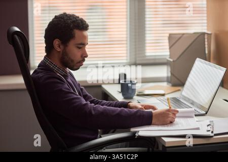 Side view of a young man at a desk in an office, reviewing documents by a window. The professional setting emphasizes desk work, business and modern workplace. Stock Photo