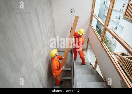 Construction workers carrying long boards up narrow stairs inside a house under construction, some wearing hard hats. The scene highlights teamwork and building trades on a construction site. Useful for industry, construction and trades photography. Stock Photo
