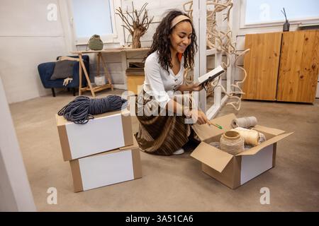 Dynamic image of a Black female entrepreneur with curly hair in boho style, packing boxes with products for delivery in an art studio. Perfect for features on women-owned businesses, handmade products, and online shops needing packaging and fulfillment visuals. Great for ecommerce marketing, small business stories, and creative branding. Stock Photo