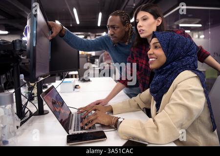 Black businesswoman wearing hijab works on a laptop at a table with coworkers standing beside her in the office. They review code and collaborate on a project, illustrating diverse tech teams and startup culture. Stock Photo