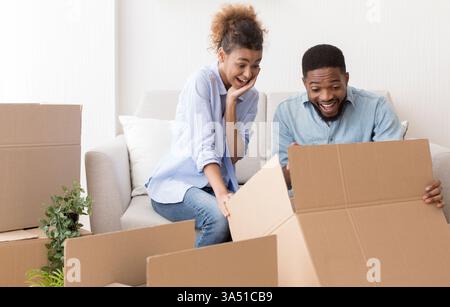 Excited African American couple unpacking moving boxes on a sofa in their new apartment. The image captures relocation and home ownership, with copy space for branding. A versatile choice for real estate, moving, or lifestyle campaigns. Stock Photo