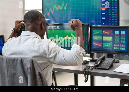 Rear view of a young Black man in formalwear gazing at a computer screen as stock charts rise and he feels excited. This image works well for finance, trading, investment, and business lifestyle campaigns. Stock Photo