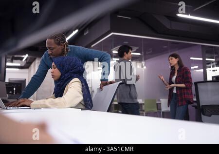 Black Muslim female software developer collaborates with a male colleague, coding on a laptop in a modern office. This diverse tech scene emphasizes teamwork and software development. Stock Photo