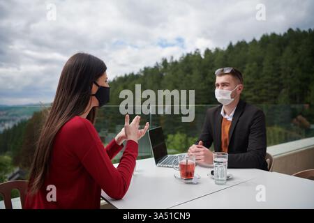 Group of casually dressed professionals outdoors, wearing protective masks while collaborating over a meal. The scene captures teamwork and safety during the pandemic era. Ideal for business, health, and hospitality contexts with a focus on collaboration. Stock Photo